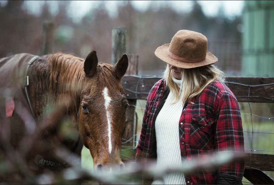 A person in warm autumn plaid and a sweater with the brim of their cat covering their eyes leads a gentle looking brown horse near the fence of the corral on the farm. 