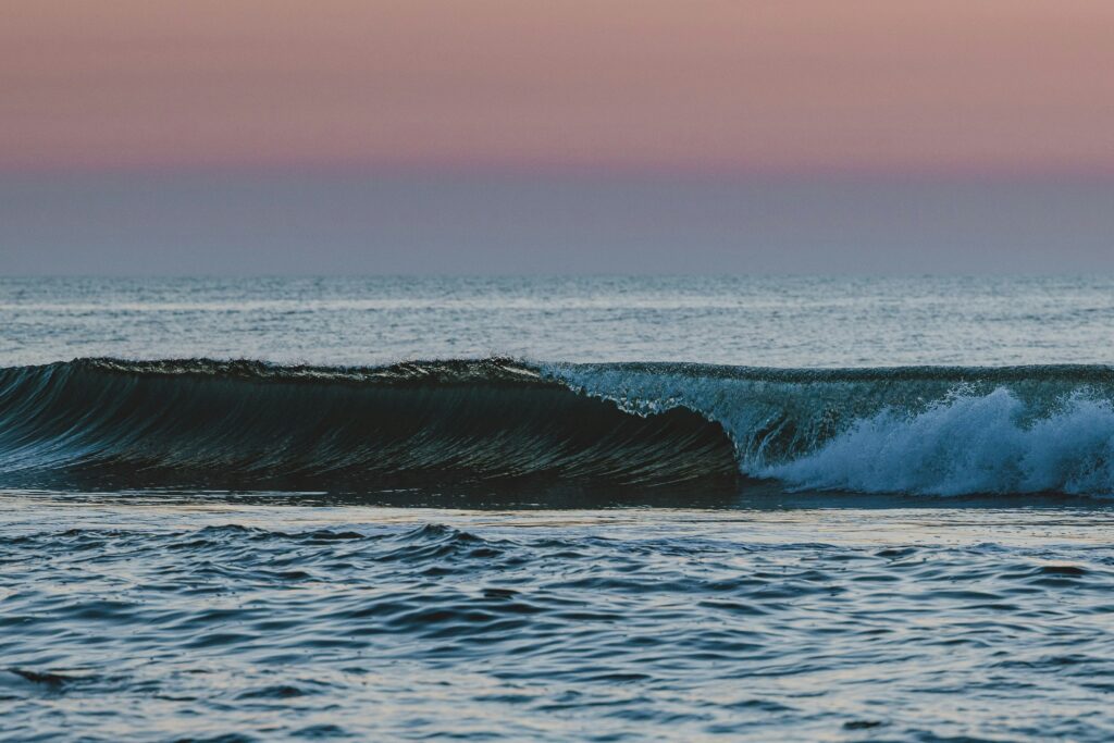 A single wave folds over and crashes with a pink sunset in the background.