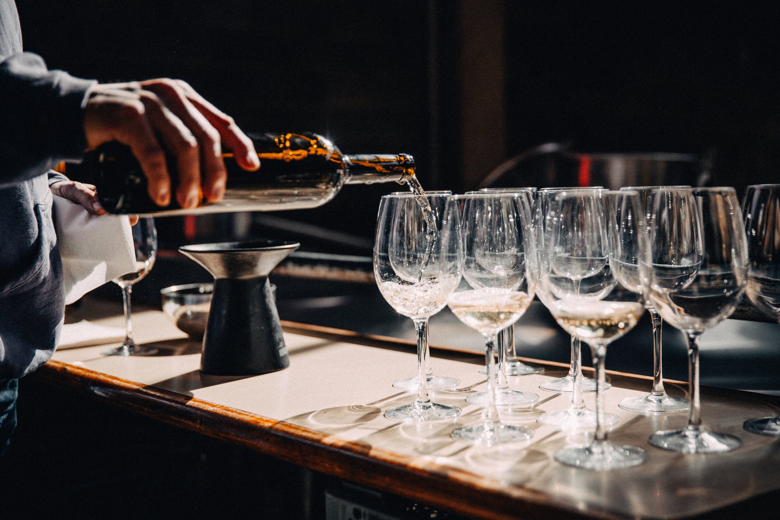 A man pours white wine into 11 glasses that sit on a wooden bar counter.