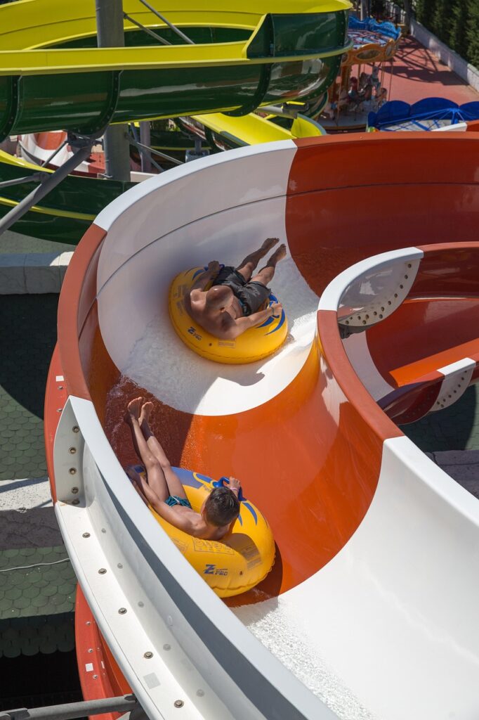 Two boys on two yellow inner tubes glide down a red and white stripped waterslide in the bright sunshine.