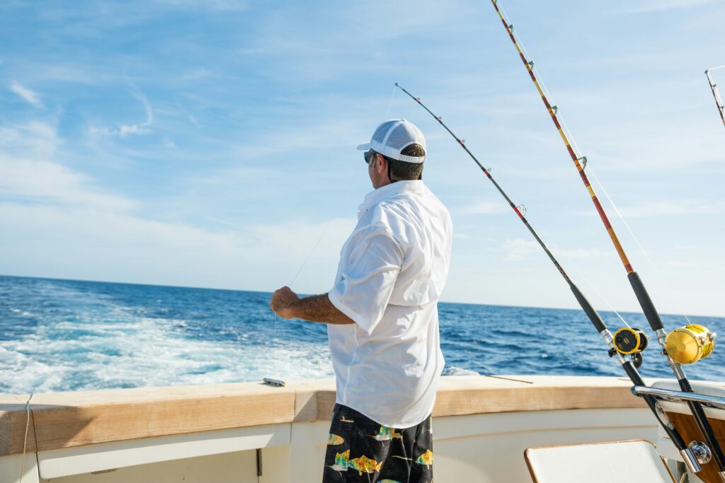 A man wearing a bathing suit and white shirt looks off the back of the boat at his two fishing poles bent into the water. 