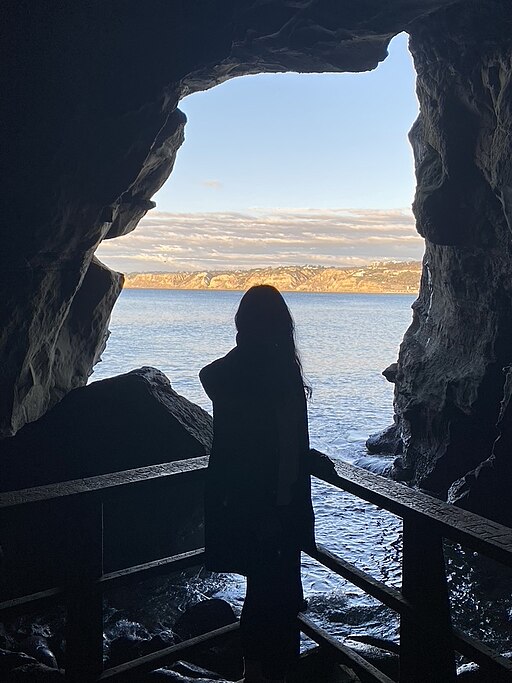 A woman stands at the opening of Sunny Jim's Cave looking out at the Pacific Ocean. The opening of the cave's silhouette matches the silhouette of the mascot of the cereal.