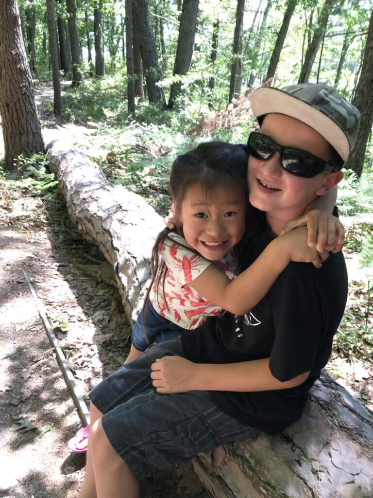 Two siblings rest on a log along a path in the woods as sunlight streams in-between the leaves. They hug each other and smile at the camera.
