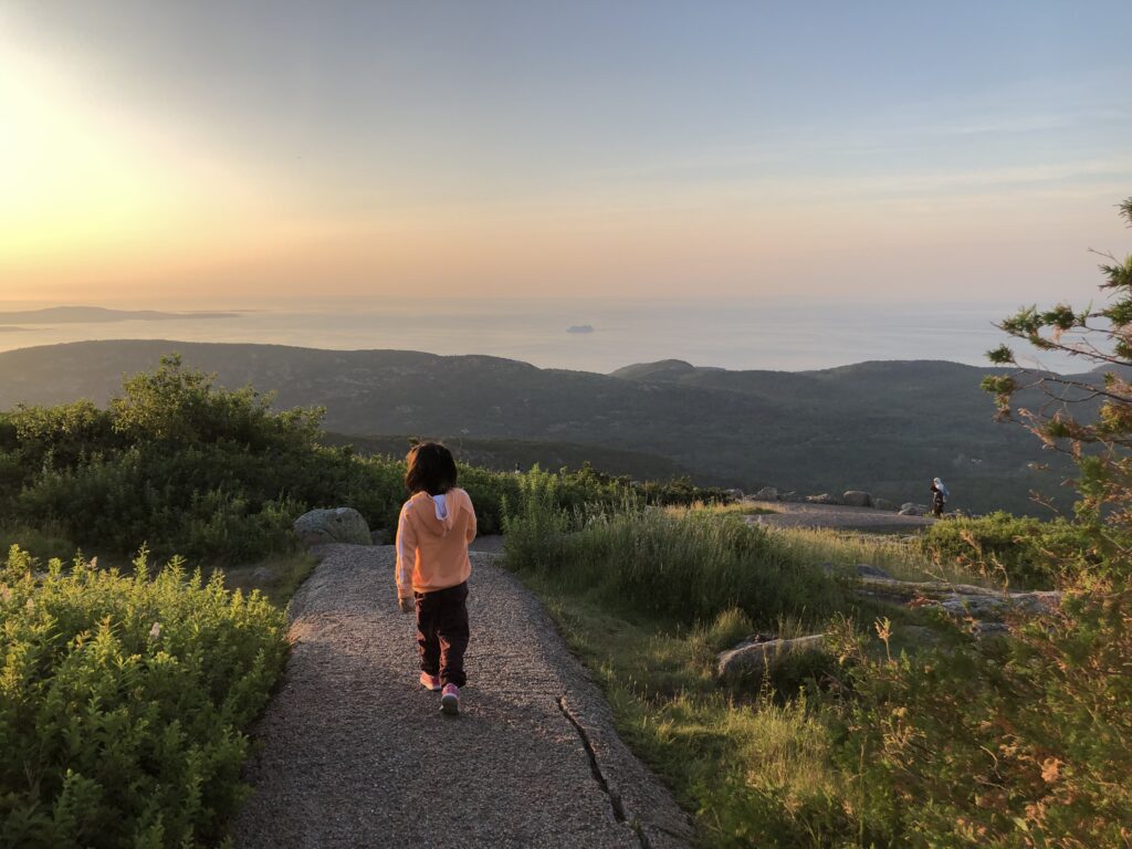 A young girl walks confidently ahead looking at the sunrise on Cadillac Mountain.
