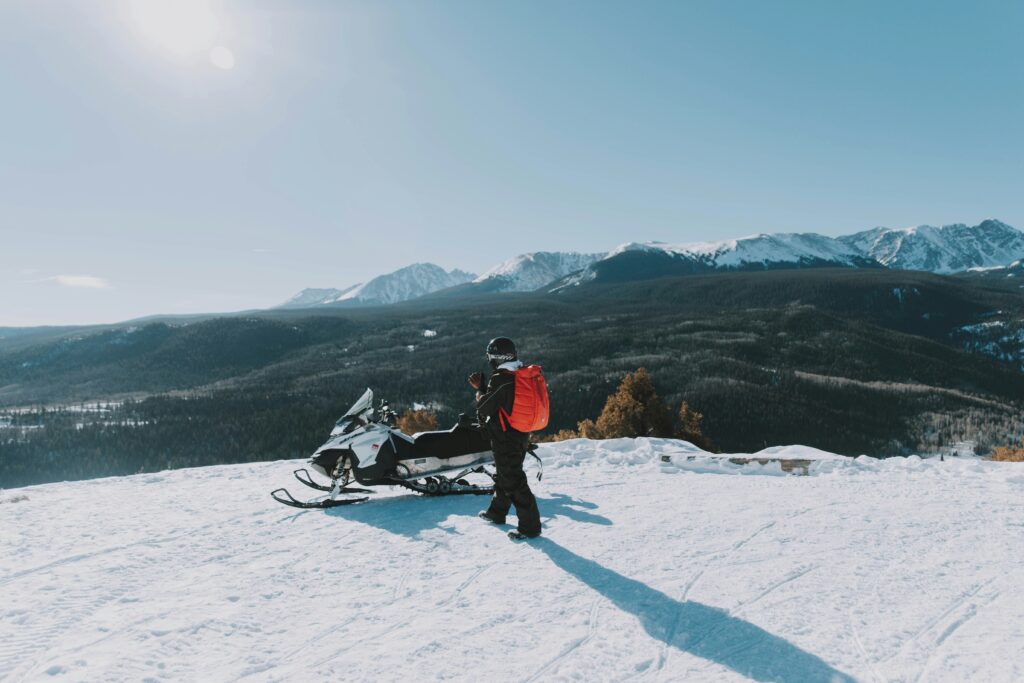 A man stands next to his snowmobile and admires the views of the snow capped mountains in the distance.