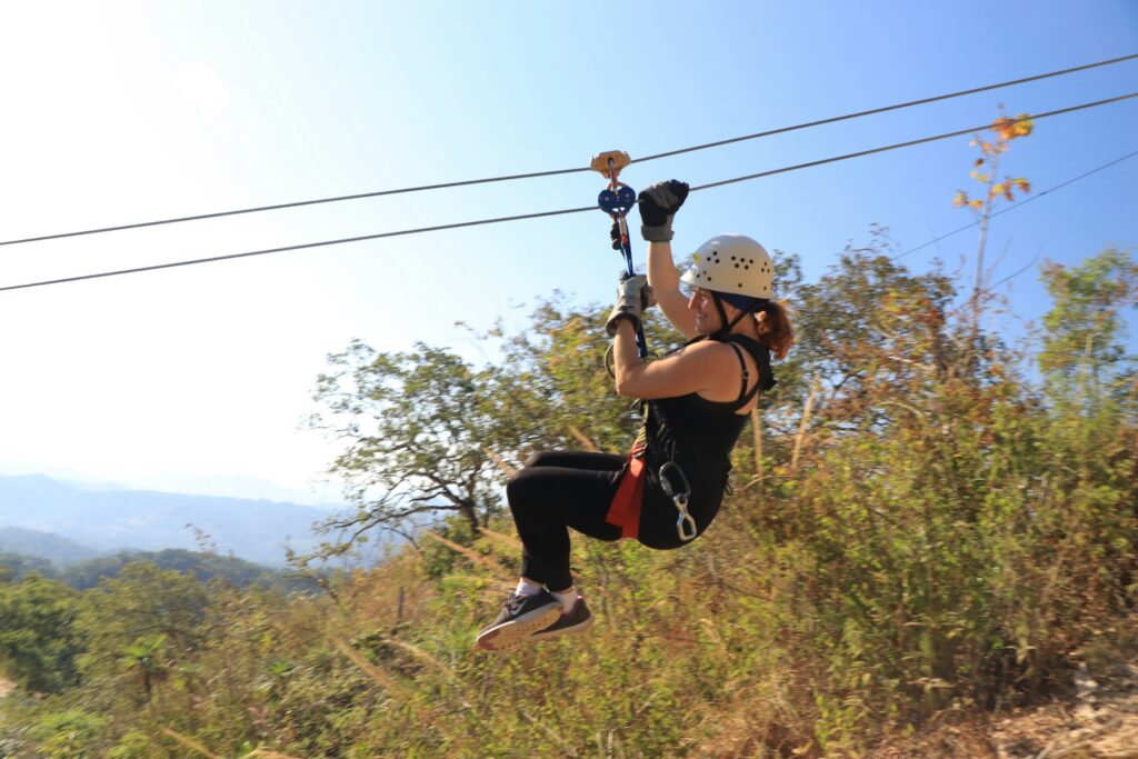 Woman in a white helmet glides cautiously down a zipline holding on to the line with thick gloves. 