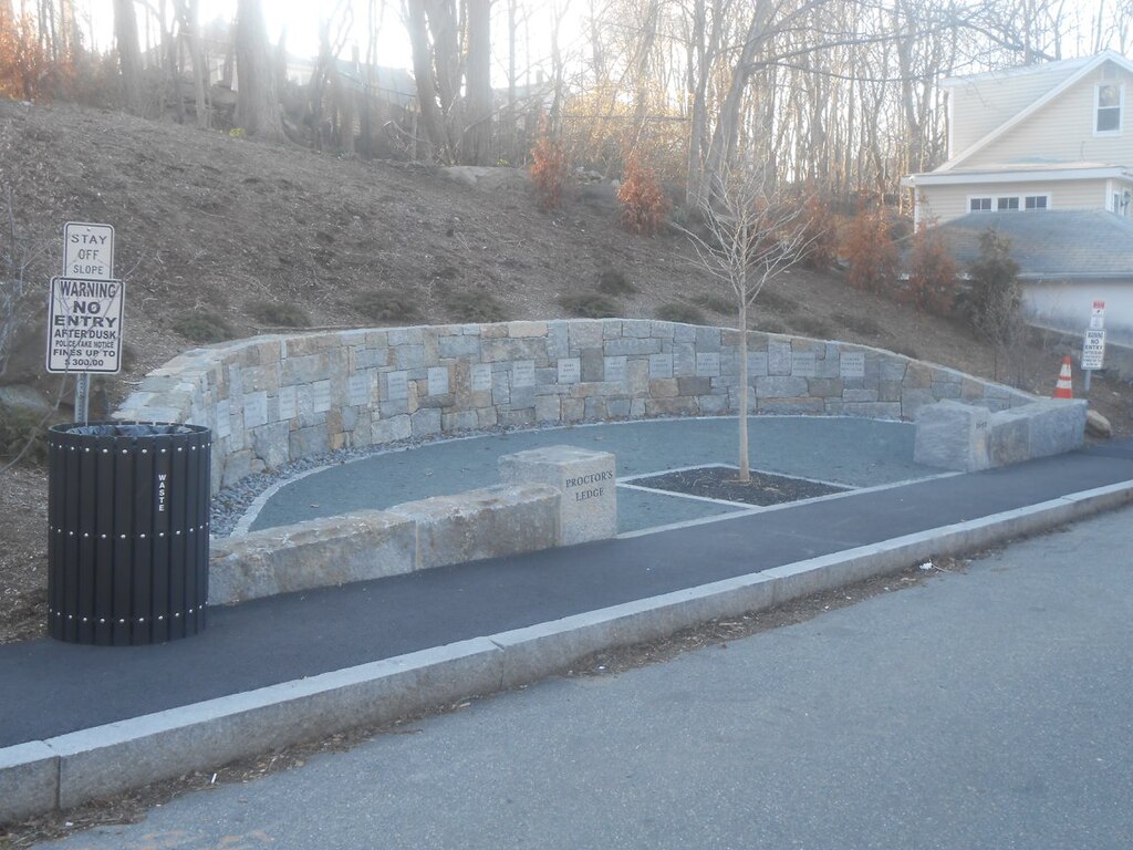 A small tree stands in front of an arched stone wall. Square stones list the names of the victims who were deemed to be witches and executed at this site. 