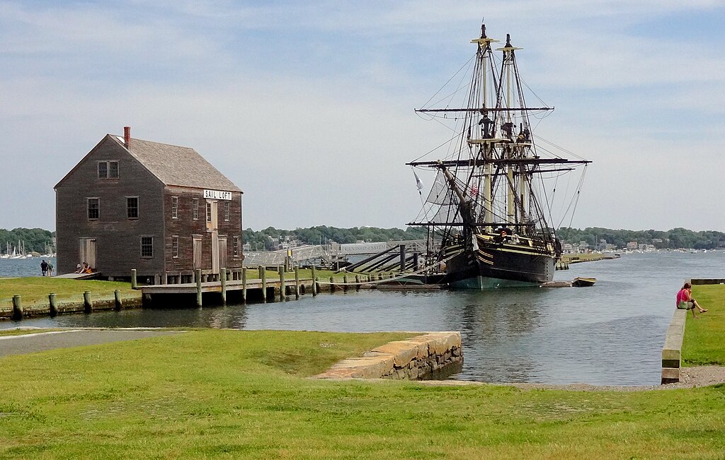 A large historic ship sits in the water near a historic house along the water's edge in Salem, Ma.